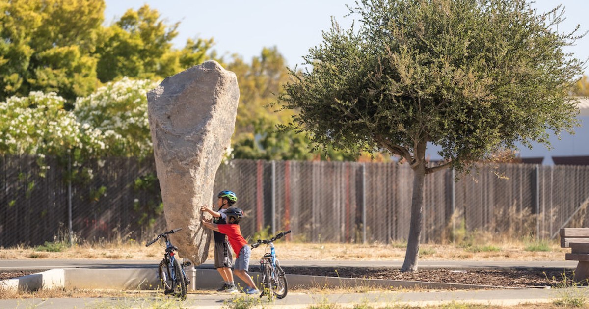 Two kids on bikes push against Oumuamua, one of the granite Rockspinner sculptures at Jean Sweeney Open Space Park in Alameda